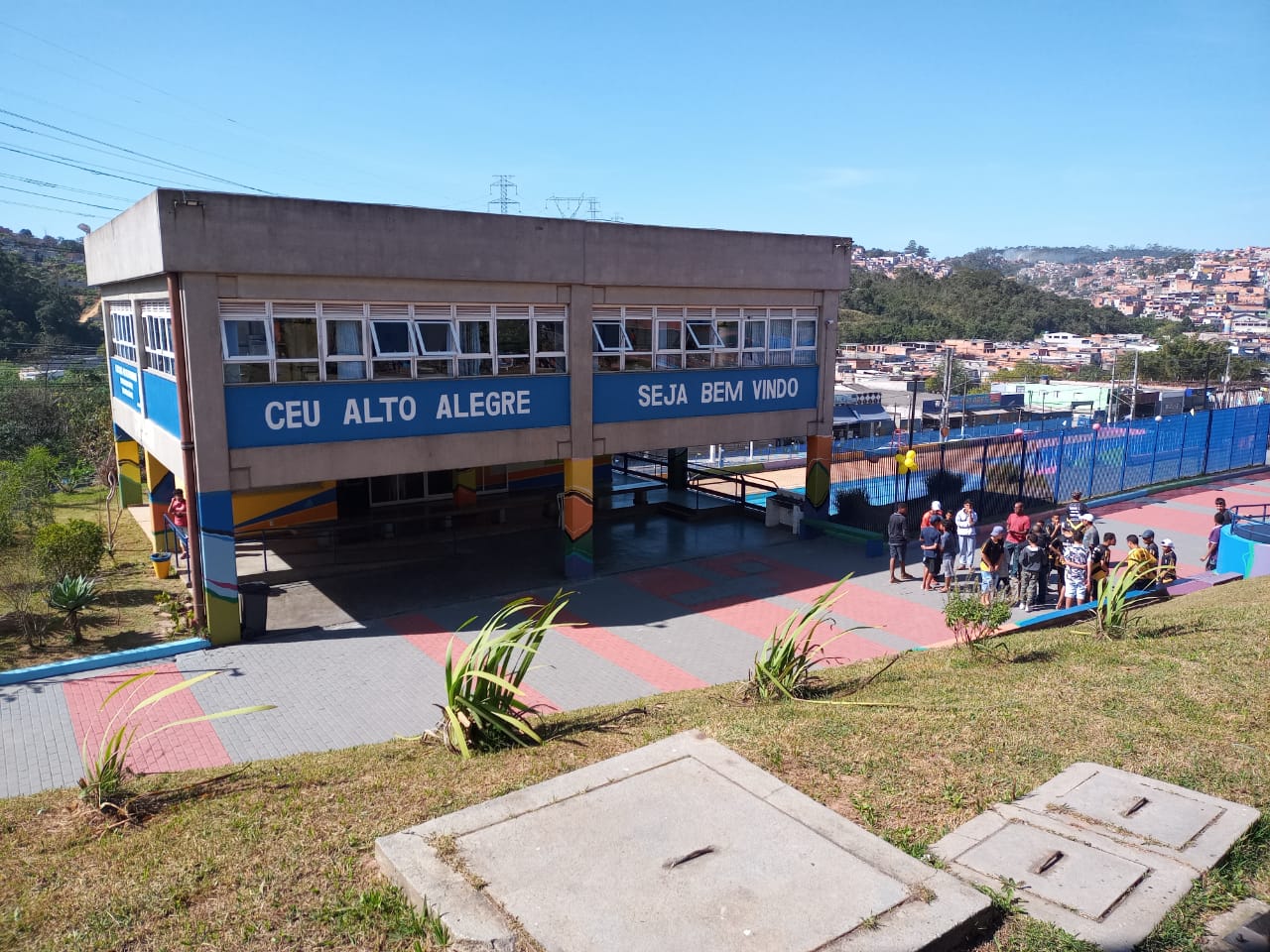 Grande prédio com janelas e uma faixa azul no centro com os dizeres "CEU ALTO ALEGRE, SEJA BEM VINDO". Ao lado direito da imagem, vemos um amontoado de pessoa